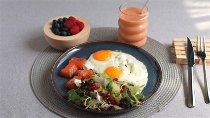 Vibrant eggs, salmon slices, mixed green salad with pomegranate seeds, orange smoothie, and fresh berries highlight balanced eco-friendly meal focused on wellness and clean eating.