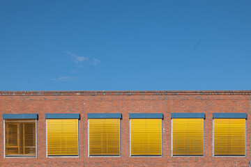 Rote Backsteinfassade mit Fenstern, gelbe Rolläden, blauer Himmel