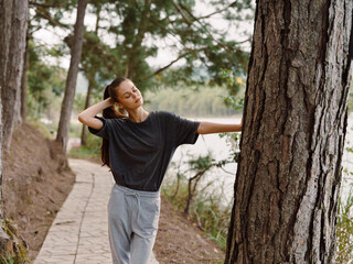 Relaxed young woman leaning against a tree by a serene lakeside path, enjoying nature and the fresh air Representing peace and tranquility in the outdoors