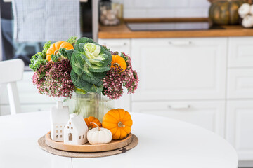 Still-life. A bouquet of decorative cabbage in a vase, pumpkins and white houses on the table in the interior of a home kitchen. A cozy autumn concept.