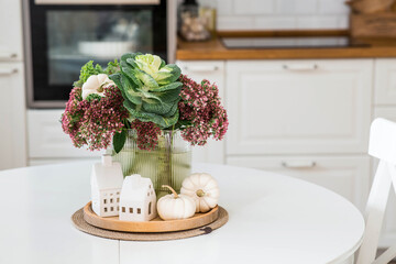 Still-life. A bouquet of decorative cabbage in a vase, pumpkins and white houses on the table in the interior of a home kitchen. A cozy autumn concept.