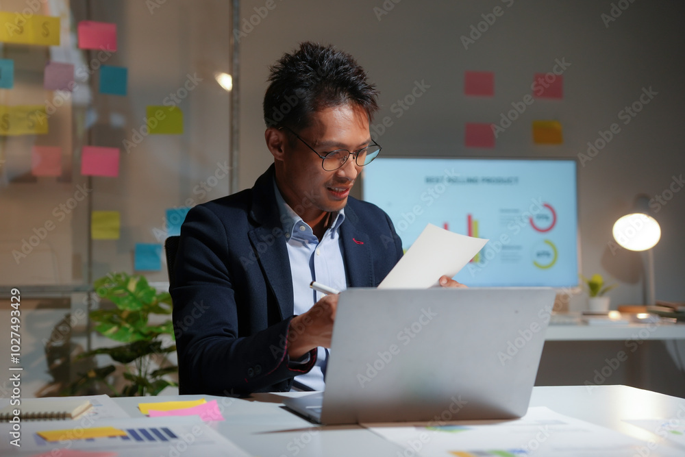 Poster smiling asian businessman working late analyzing documents and using laptop computer in modern offic