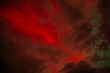 A dramatic view of the Aurora Borealis with intense red hues glowing through a cloudy night sky. The vibrant red contrasts with the dark clouds, creating a fiery and mysterious atmosphere.