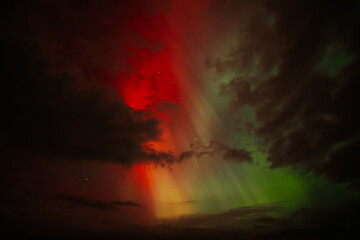 Fototapeta premium A vivid display of the Aurora Borealis lights up the night sky, with striking red, orange, and green hues blending amidst dark clouds. The dramatic contrast creates a surreal and captivating scene.