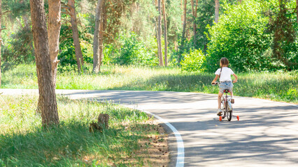 Fototapeta premium Girl cyclist in park leads an active lifestyle outdoors. Kid dreams of traveling by bike. Child rides a bicycle on road in the forest. Girl in the park in nature rides a bicycle on the road