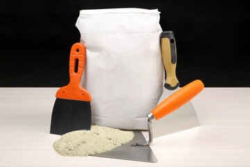 Bag of cement powder and putty knives on light wooden table against black background. Building...