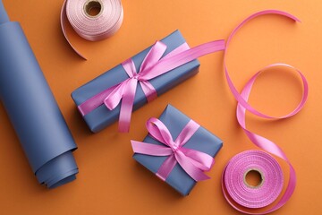 Beautiful gift boxes with pink bows, ribbons and roll of wrapping paper on orange background, flat lay