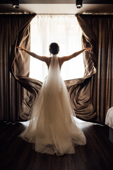 Fototapeta premium The girl stands with her back near the window and opens the curtains with her hands