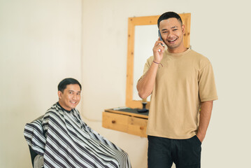 A man is engaged in a conversation on his cell phone beside another man who is receiving a haircut at a nearby salon or barbershop