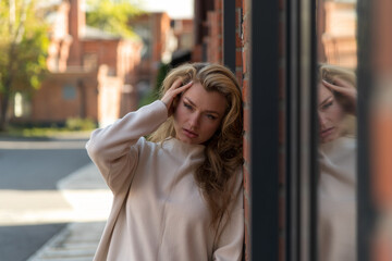 Fototapeta premium Close-up portrait of beautiful serene long haired blond woman in white sweater standing outdoor on city street by red brick loft building wall and reflects in window in autumn day. Fashion theme.