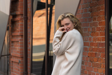 Fototapeta premium Portrait of beautiful happy long haired caucasian blond woman in white sweater with handbag walking or standing outdoors on city street by red brick loft building. Soft focus. Fashion and beauty theme