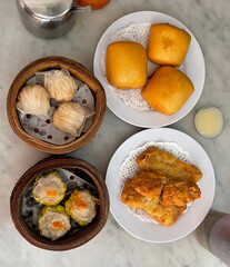 Top-down view of four plates of freshly served dim sum on a marble table. Perfect for food blogs, menus, and Asian cuisine promotions