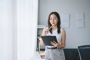 Young manager woman is thinking about a challenging task while holding a clipboard and a pen in her office