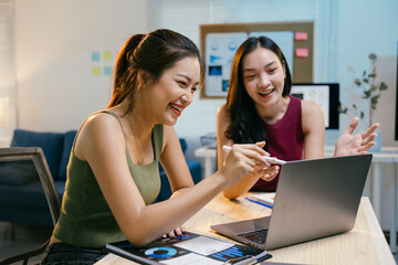 Two young businesswomen are enjoying themselves while working together on a laptop in their office. They are looking at the laptop screen and laughing