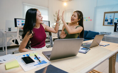 Two businesswomen celebrate success with a high-five in a busy office, surrounded by laptops and paperwork