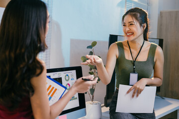 Two young businesswomen discussing financial data in a modern office, using a computer and documents, one smiling and gesturing while explaining