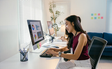 Young asian businesswoman is smiling while typing on a keyboard and working on a desktop computer. She is sitting at her desk in a modern office