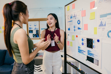 Two young businesswomen are discussing new ideas in front of a whiteboard full of charts and...