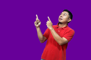 An excited Asian man wearing a red shirt points to the empty space above him, isolated on a purple background.