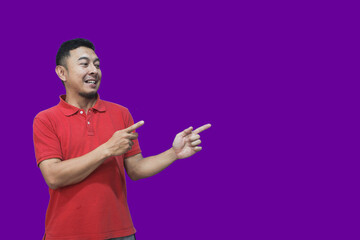 An excited Asian man wearing a red shirt points to the empty space above him, isolated on a purple background.