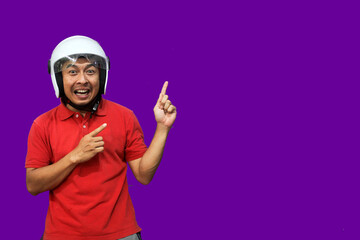 Young Asian driver man wearing helmet looking up while raising both hands and pointing upwards with happy expression
