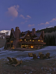 A rustic lodge at dusk, surrounded by snow and mountains, illuminated warmly.