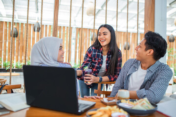 Obraz premium Three young Indonesians use a laptop while relaxing and chatting in a coffee shop
