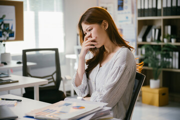 Obraz premium Young businesswoman is sitting at her desk with her hand over her mouth, feeling the stress of working with overwhelming paperwork