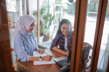 seen from the window two Asian women sitting working on a laptop together in a coffee shop