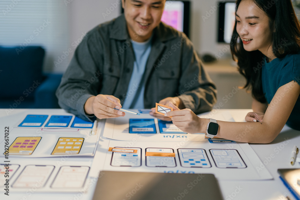 Leinwandbilder Two asian ui/ux designers are working together in office, holding ui element icons and discussing about mobile app interface wireframe design on table