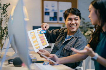 Two asian ui/ux designers collaborating on a mobile app wireframe in an office late at night, brainstorming and strategizing to bring their creative vision to life
