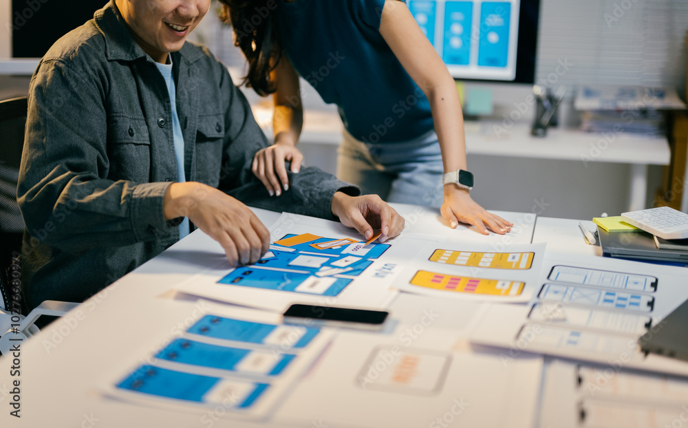 Poster Two designers collaborate on a mobile app wireframe, discussing ideas for a user-friendly interface. The vibrant office reflects their teamwork and creativity