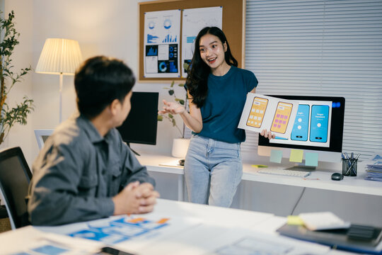 Two asian ui/ux designers collaborating on a new mobile app, discussing interface design in an office filled with computers