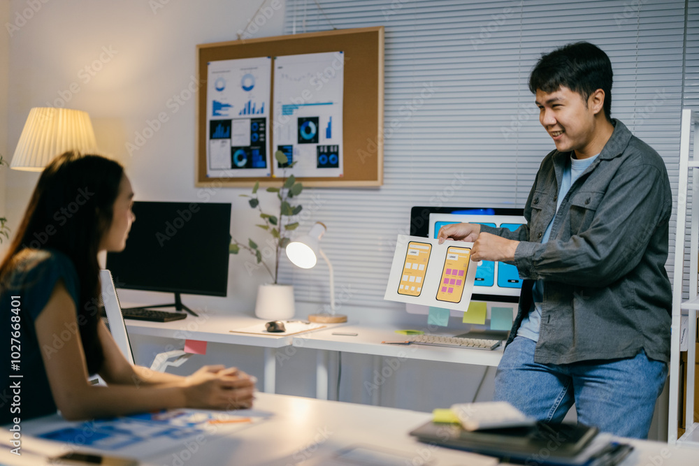 Poster two asian ui/ux designers collaborate late at night in an office. discussing mobile app interface de
