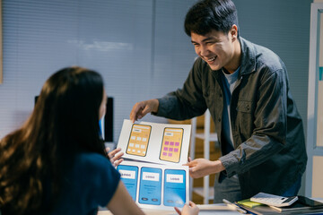 Two asian ui/ux developers are discussing new mobile application wireframe designs, working late at...