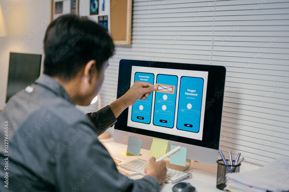 Poster Ui developer is working on a user interface for a mobile application, using a wireframe kit displayed on his computer screen. He is holding a pen in his other hand and seems to be taking notes