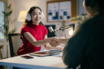 Obraz premium Two happy radio hosts shaking hands while recording podcast in home studio. They are sitting at desk with microphone and headphones