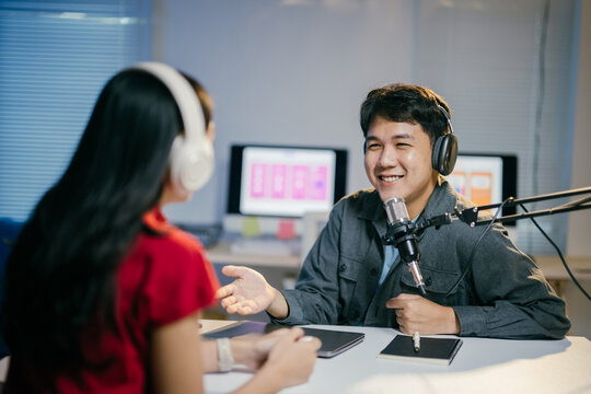 Two young asian radio hosts record a podcast in a vibrant studio. Discussing various topics with teamwork and camaraderie. Smiling and immersed in technology. They create entertaining online content