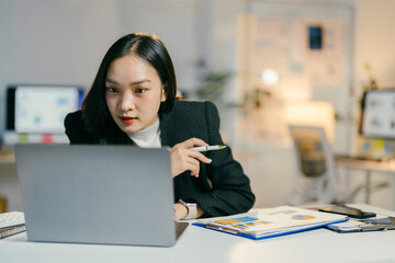 Fototapeta premium Focused asian businesswoman works late at night in her office, analyzing data and preparing for a presentation. She is determined and ambitious, showcasing her dedication to her career
