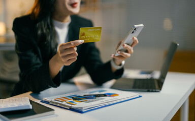 Focused businesswoman multitasks at her desk, making an online payment with a credit card and smartphone, showcasing the seamless integration of technology in modern business practices