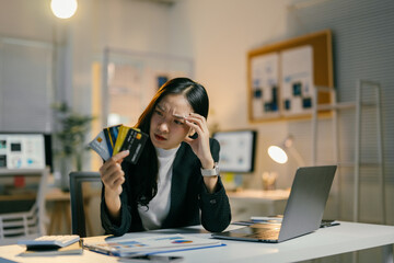 Young professional is sitting at her desk, stressed out and holding multiple credit cards. She is...