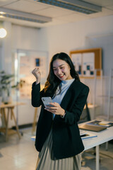 Young businesswoman celebrates success in her office, holding a smartphone and raising her fist in victory, exuding excitement and happiness