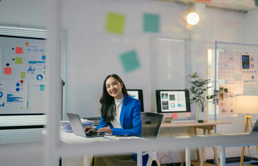 Businesswoman is sitting at her desk working on her laptop in a modern office setting with graphs...
