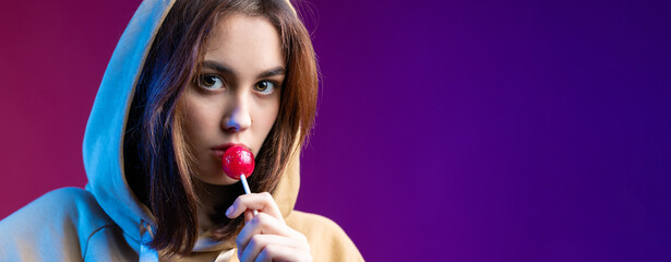 Portrait of a woman in a hoodie, girls licking a red round lollipop with beautiful makeup on a purple trendy background in the studio.