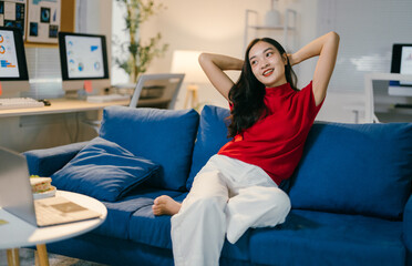 Obraz premium Young asian woman is relaxing on her couch at her home office after a long day of work, with her laptop and a snack on the table in front of her