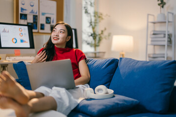 Obraz premium Young asian woman relaxes on a blue sofa in her home office, working on her laptop with her feet up, smiling and enjoying the flexibility of working from home
