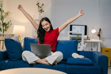 Young asian woman is sitting on her sofa with her laptop, she is raising her arms in the air with a...