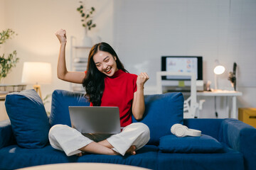 Young asian woman happily works on her laptop at home, celebrating success with a fist pump in her...