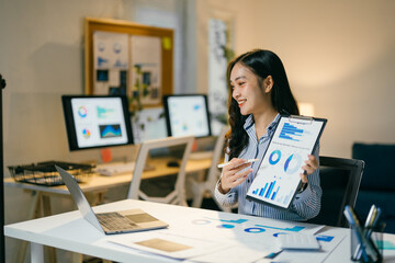 Smiling asian businesswoman presenting charts and graphs on clipboard while having video call on laptop with colleagues