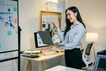 Asian businesswoman is holding a laptop and making a fist pump, excited about a recent business...